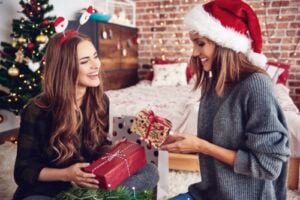Two women exchanging wrapped presents in a bedroom, one in a Santa hat, Christmas tree behind