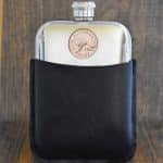 1938 Australian penny hip flask in a black leather sleeve, kangaroo coin set into the polished pewter front, dark wood backdrop