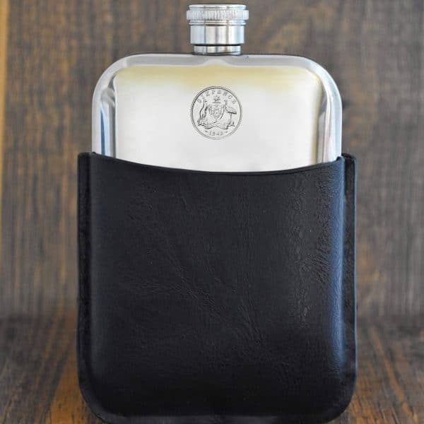 1943 Australian sixpence hip flask, coat of arms coin set into the polished silver-tone front, black leather sleeve, dark wood backdrop
