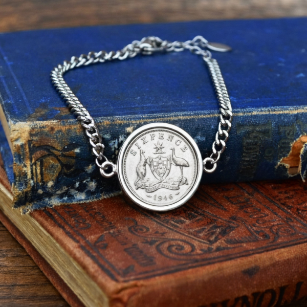 1946 Australian sixpence in a silver-tone bezel on a curb-chain bracelet, coat of arms reverse, resting on old blue and red books