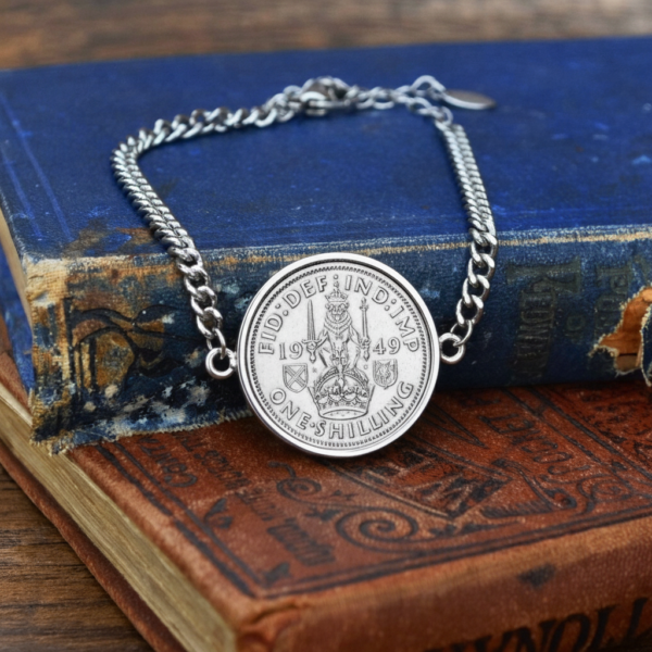 1949 one shilling coin in a silver-coloured bezel on a curb chain bracelet, Scottish lion and crown reverse, resting on old cloth-bound books
