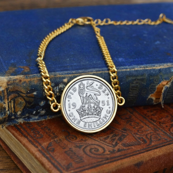 1951 one shilling coin in a gold-coloured bezel on a gold curb chain bracelet, laid on worn antique books
