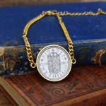 1959 one shilling coin in a gold-coloured bezel on a gold curb chain bracelet, resting on old blue and brown books