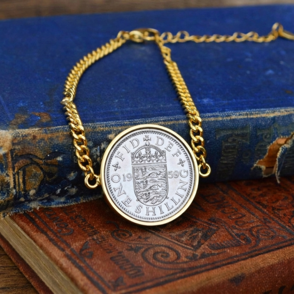 1959 one shilling coin in a gold-coloured bezel on a gold curb chain bracelet, resting on old blue and brown books