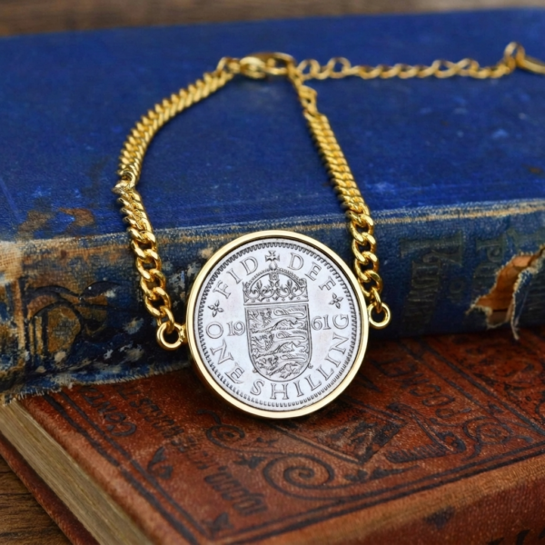 1961 one shilling coin in a gold-coloured bezel on a gold curb chain bracelet, resting on old blue and brown books