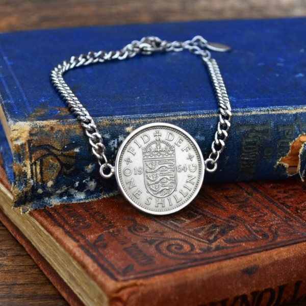 1964 one shilling coin bracelet on a silver-coloured curb chain, English shield side up, laid on worn blue and red books