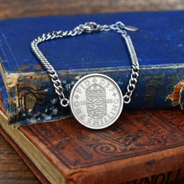 1966 one shilling coin bracelet on a silver-coloured curb chain, English shield side up, laid on worn blue and red books