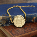 1966 threepence coin bracelet on a gold-coloured curb chain, twelve-sided coin with crowned portcullis, on old books