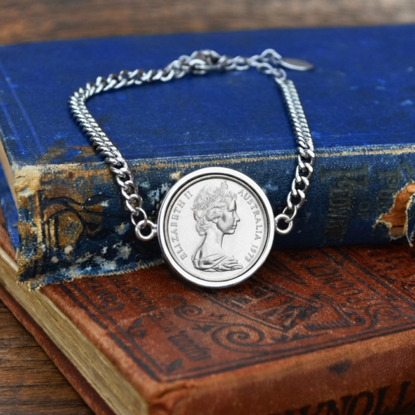 1972 Australian coin in a silver-tone bezel on a silver-tone curb chain bracelet, laid over worn blue and red antique books