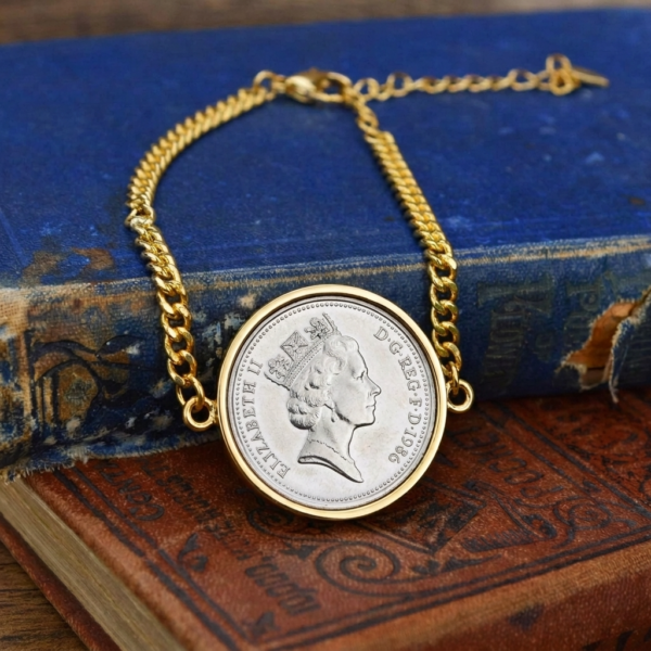 1986 old five pence coin in a gold-coloured bezel on a curb chain bracelet, laid across worn blue and red hardback books