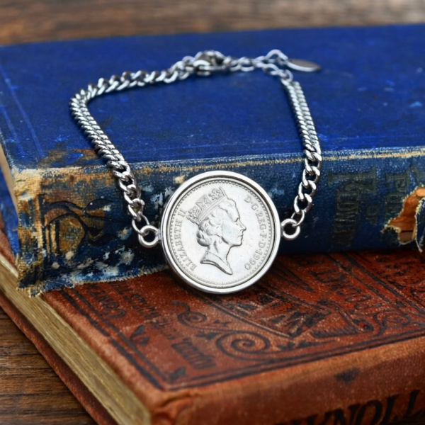 Silver-tone curb chain bracelet holding a 1990 coin, crowned Elizabeth II portrait facing right, on a worn blue book