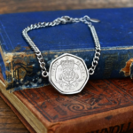 Silver-coloured curb chain bracelet holding a 1998 twenty pence coin, Tudor rose side up, on worn blue and tan books